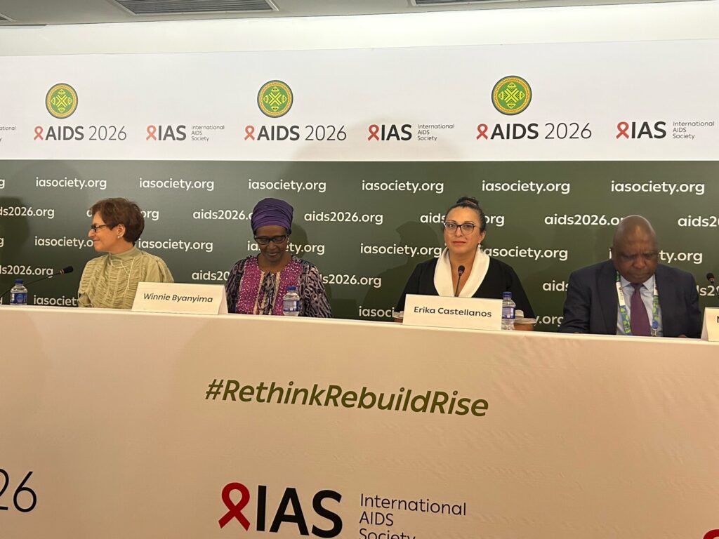 Panel of four speakers at a AIDS 2026 conference, seated behind a banner reading AIDS 2026 and iasociety.org with visible nameplates (Winnie Byanyima, Erika Castellanos).
