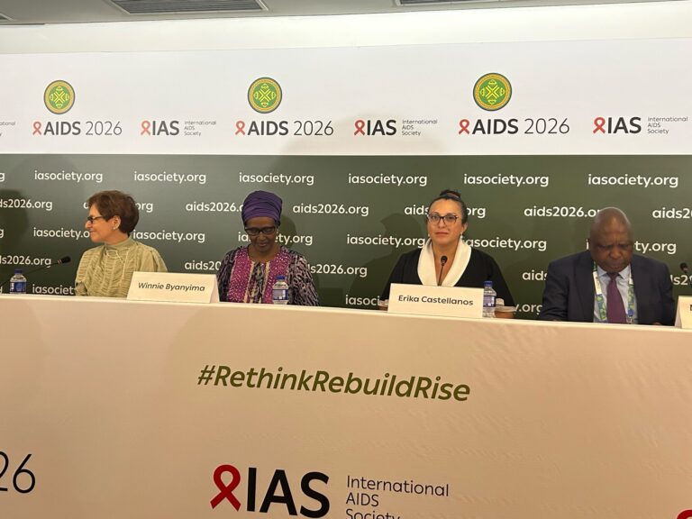 Panel of four speakers at a AIDS 2026 conference, seated behind a banner reading AIDS 2026 and iasociety.org with visible nameplates (Winnie Byanyima, Erika Castellanos).