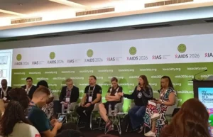 Panel discussion at the AIDS 2026 conference with six speakers seated on stage in front of a green-and-white backdrop that reads iyasociety.org and AIDS 2026