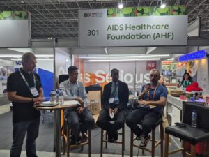 Panel discussion at a trade show booth for AIDS Healthcare Foundation (AHF), booth number 301, with four men seated and one speaking into a microphone.