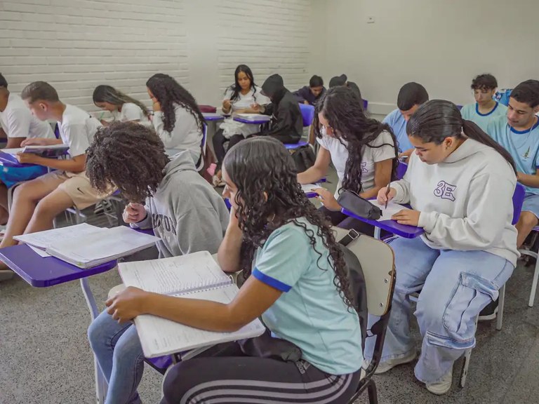 Students in a classroom seated at purple desks, writing in notebooks and studying.