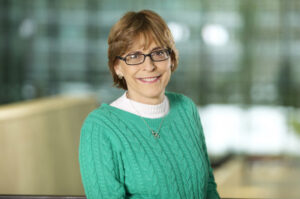 Professional headshot: smiling woman with short brown hair and glasses, wearing a green knit sweater in an office setting.