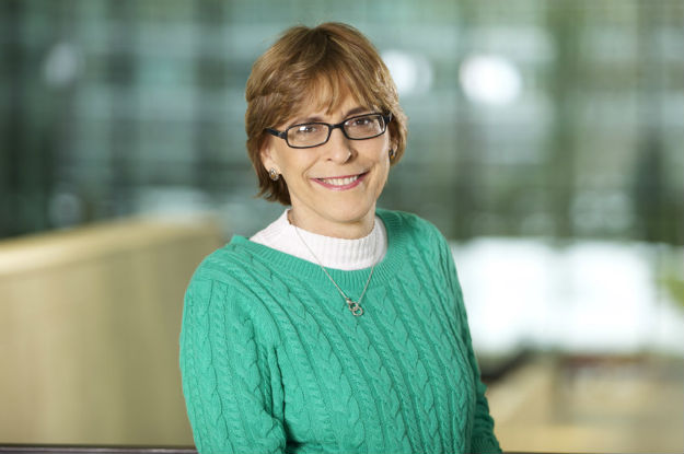 Professional headshot: smiling woman with short brown hair and glasses, wearing a green knit sweater in an office setting.