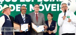 Five people on a stage during an award ceremony; a man on the right speaks into a microphone while the others hold certificates and folders.