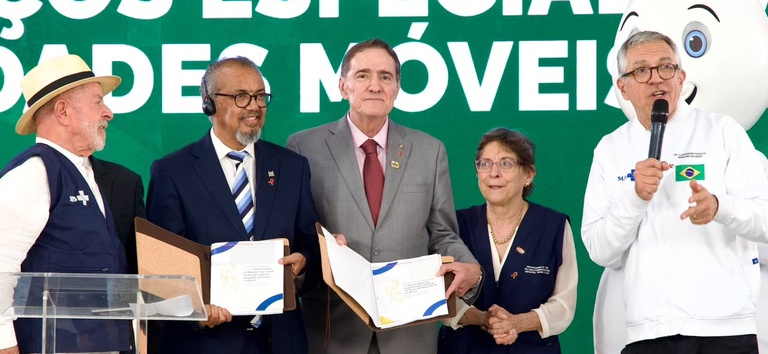 Five people on a stage during an award ceremony; a man on the right speaks into a microphone while the others hold certificates and folders.